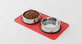 Red silicone mat with two stainless steel pet bowls one filled with kibble and the other empty.